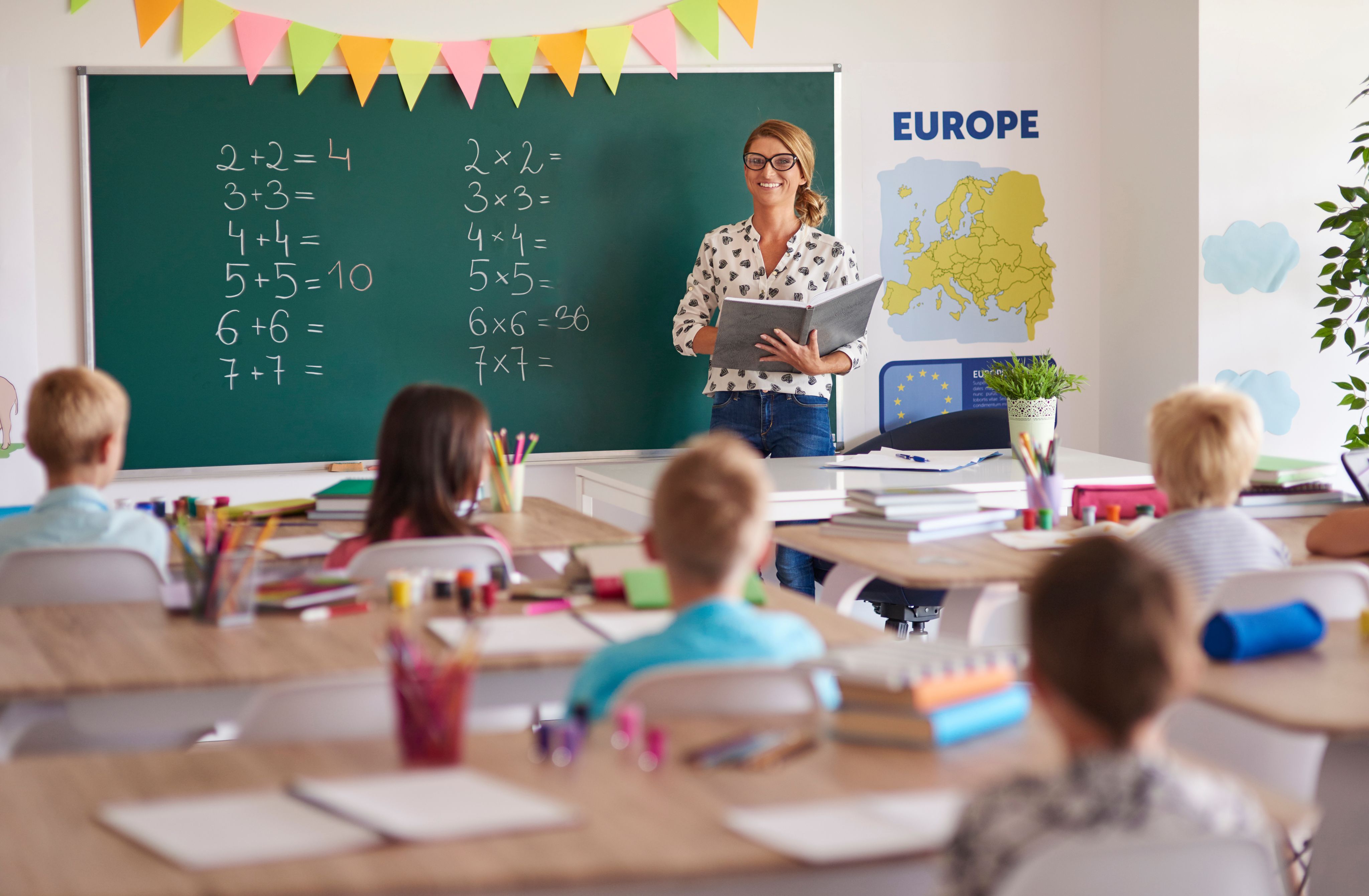 Teacher standing in front of elementary school class. Math problems are on the board and she is holding a notebook.