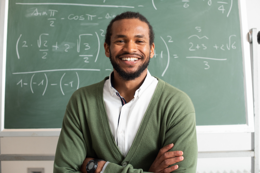 Male teacher in front of a chalboard, smiling. Math equations are written on the board. 