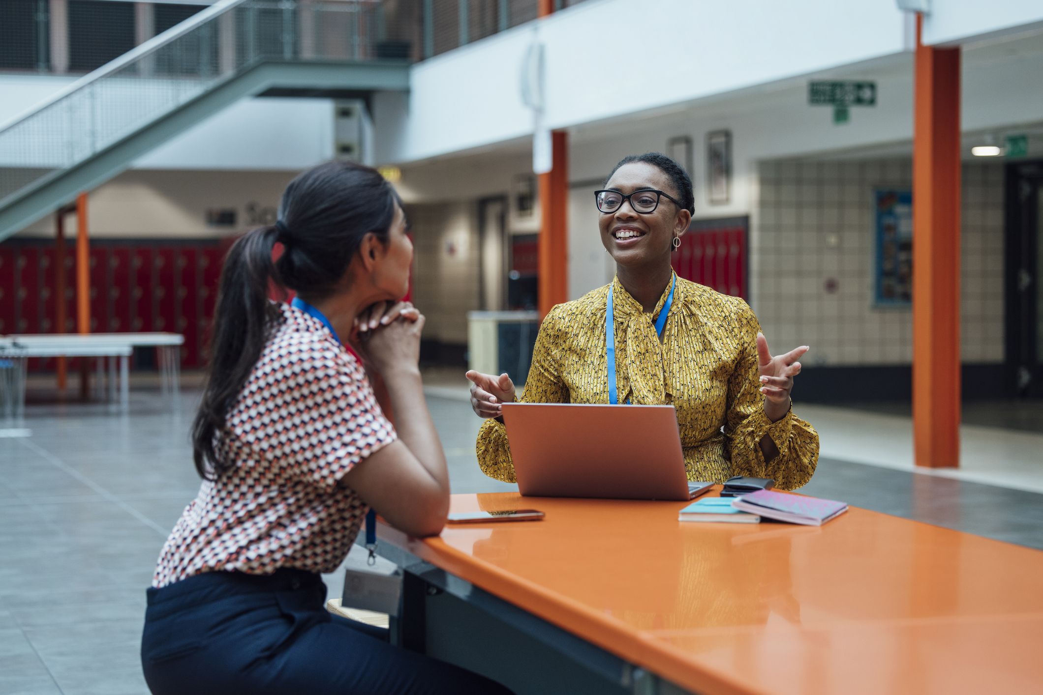 Two teachers seated at a table in cafeteria. One has a laptop and is gesturing with her hands. 