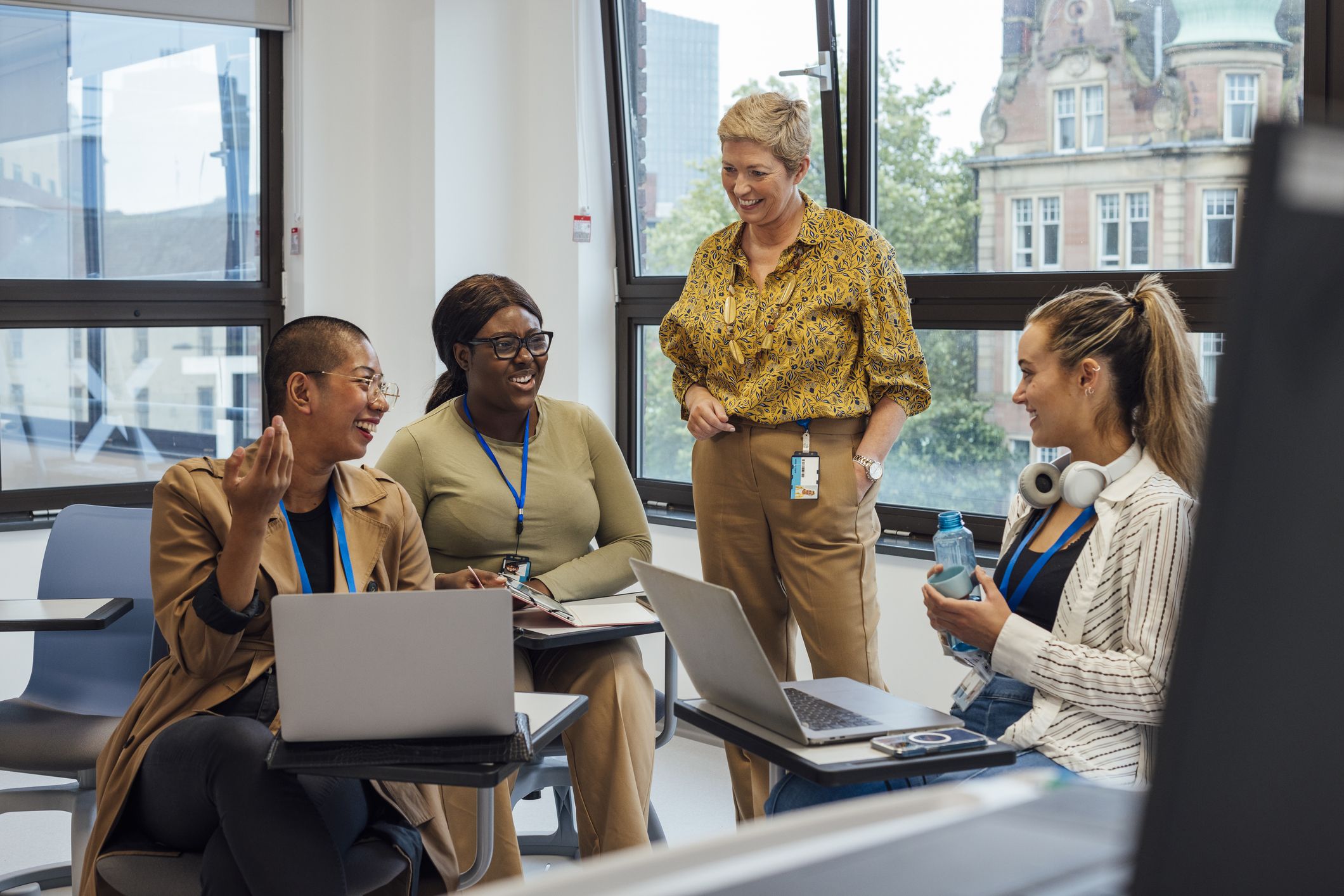 Photo of three adults at desks with laptops and a teacher or instructor standing next to them. 